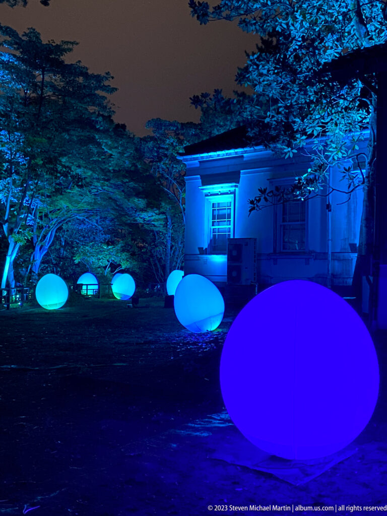 Large blue glowing responsive ovoid form with smaller lights before a blue-lit historic building.