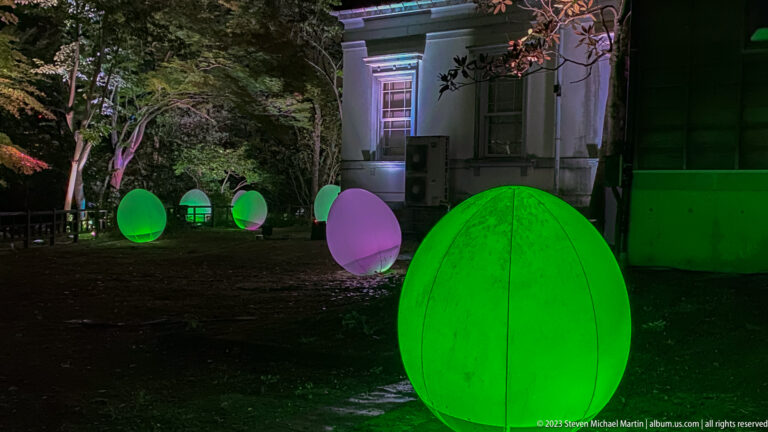 Large green glowing responsive ovoid form with smaller pink and green lights beside a building.
