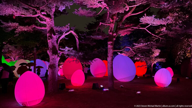 Visitors walk among multicolored responsive glowing ovoids beneath pink-lit trees.