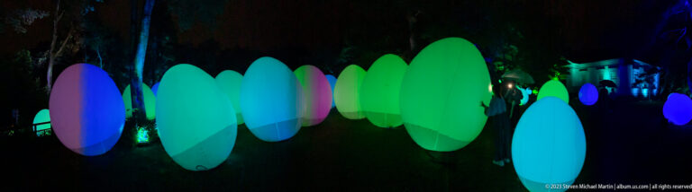 Row of blue-green glowing responsive ovoid forms leading through a dark garden toward a lit building.