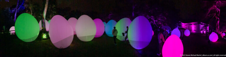 Visitors move among green, white, violet, and pink responsive glowing ovoids along a garden path.