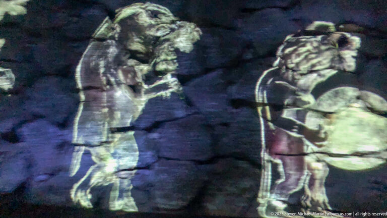Two projected costumed figures, one holding a round drum-like object, on dark stone.