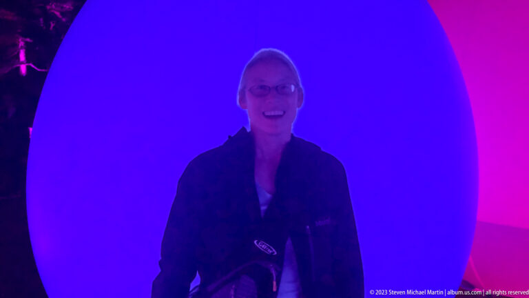 Smiling woman in dark clothing standing before large blue and pink responsive glowing ovoids.