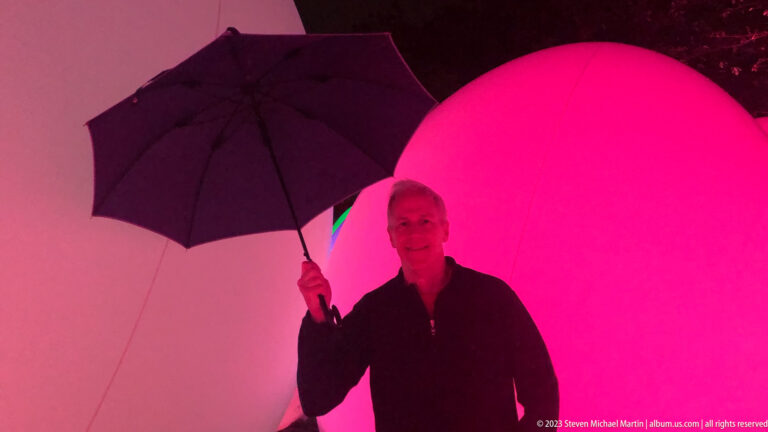 Man holding an open umbrella before large white and pink responsive glowing ovoids.