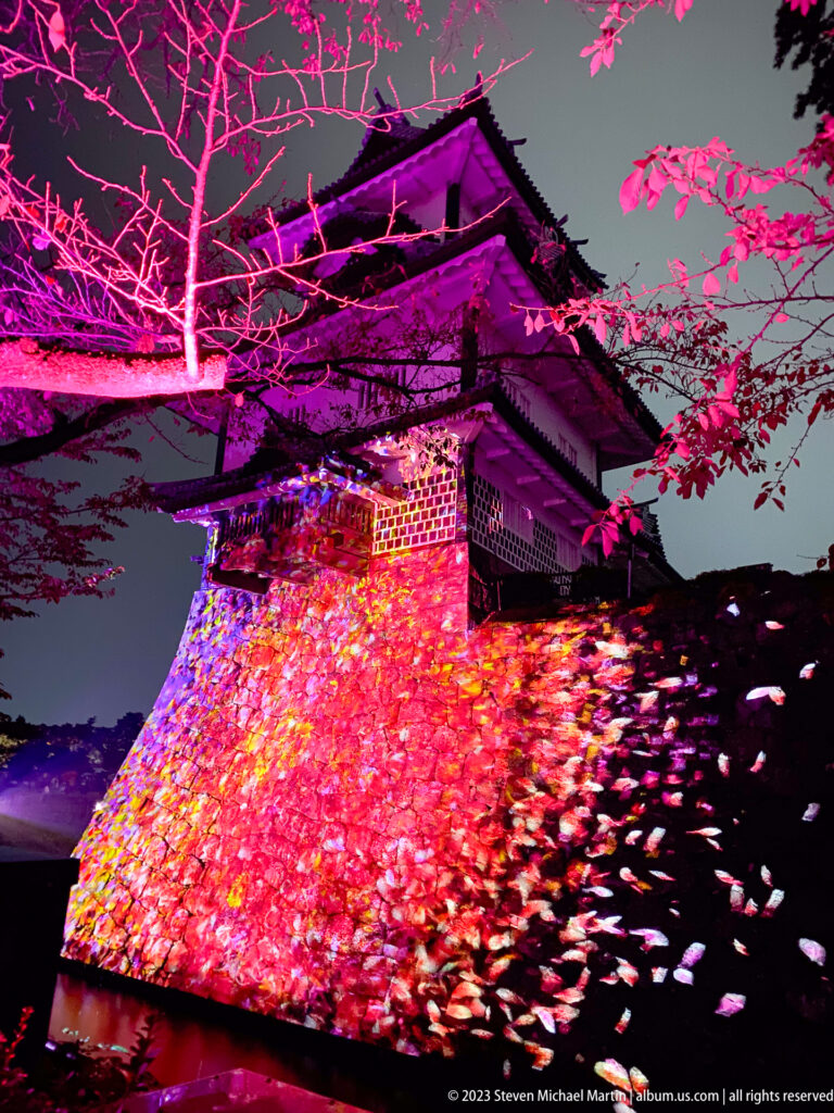 Kanazawa Castle turret above a red-pink cascade of teamLab flower-animal projections on the stone wall.