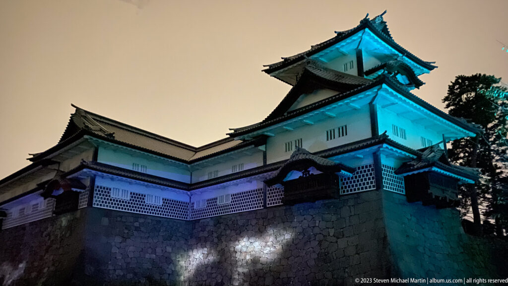 Kanazawa Castle turret and white walls illuminated blue above a high stone base at night.