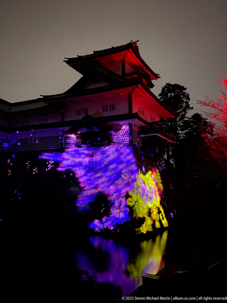 Red-lit Kanazawa Castle above colorful projections and reflections on the stone wall and water.