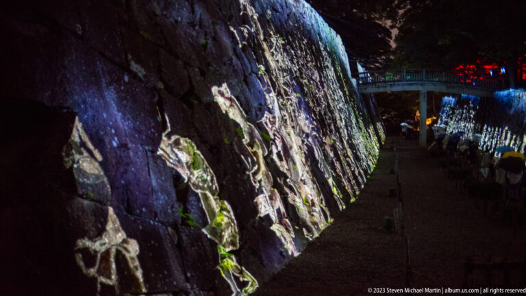 Projected human figures stretch along a rough stone wall beside a dark walkway.