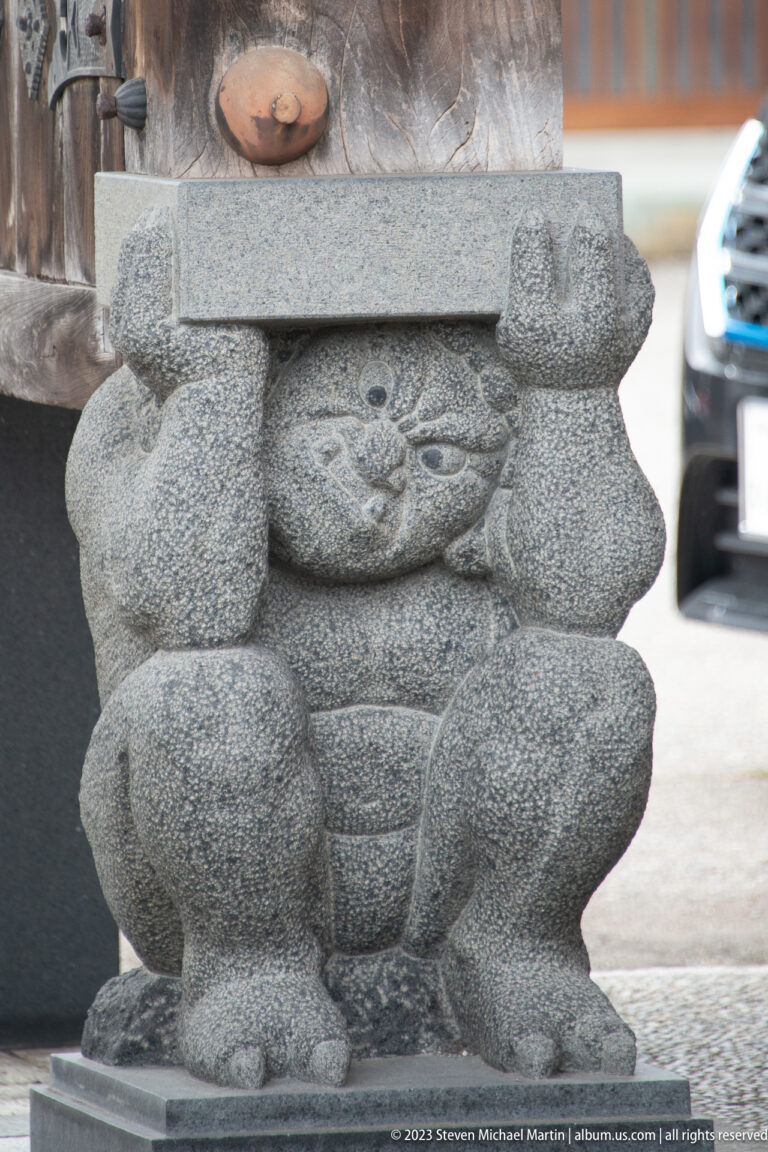 Wider view of a crouching carved stone figure beneath a monument, with pavement and street behind.