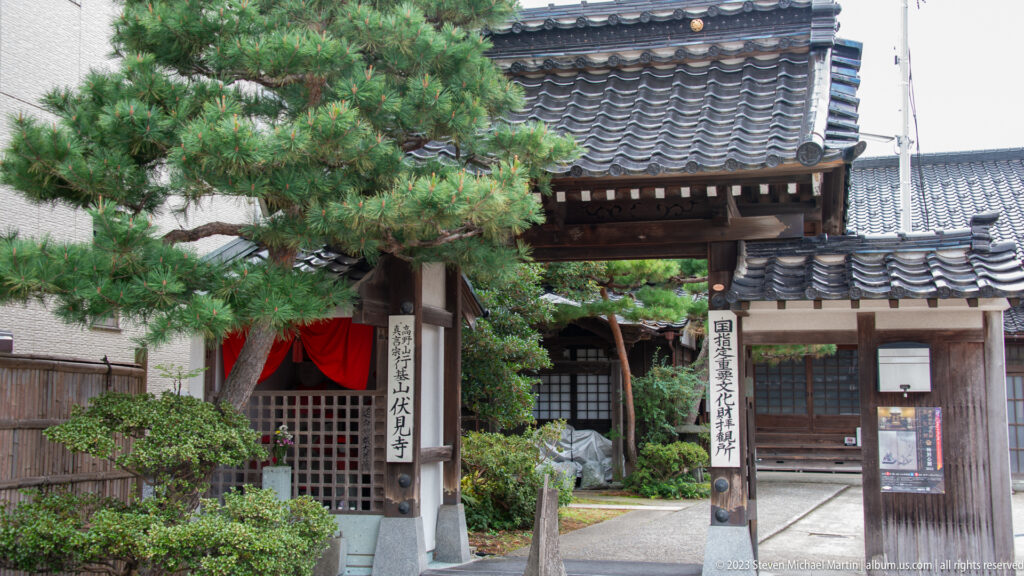 Small tiled temple gate with vertical plaques, pine tree, and red-curtained worship building behind.