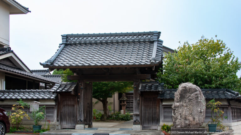 Timber gate with gray tiled roof opening into a walled compound beside a rough standing stone.