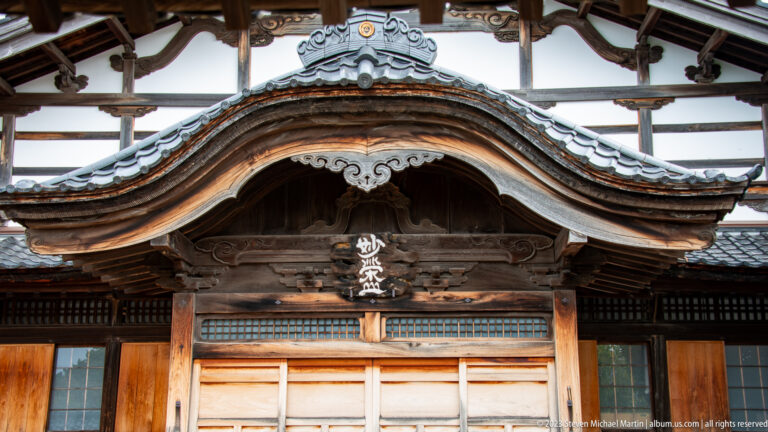 Close frontal view of an ornate curved wooden temple gable with roof tiles, carvings, and central plaque.