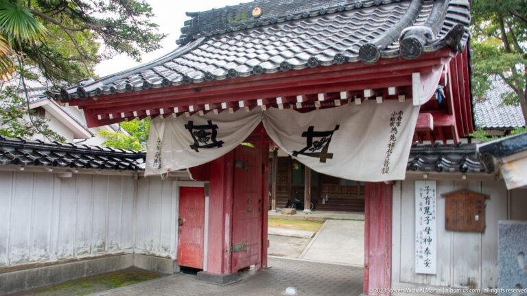 Red-painted temple gate with white emblem curtains, tiled roof, inscriptions, and greenery.