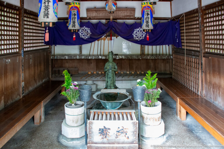 Covered wooden devotional station with central figure, basin, flowers, vessels, purple hangings, and side benches.