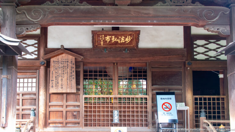 Frontal detail of a wooden temple entrance with lattice doors, carved lintel, plaque, notices, and brackets.