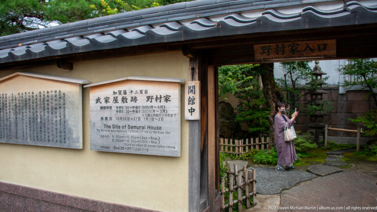 Entrance to the Nomura Samurai House with tiled roof, visitor signs, stone path, bamboo edging, and garden beyond.