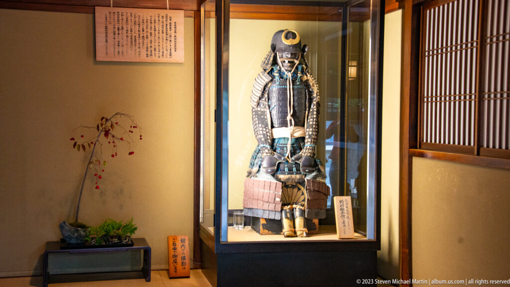 Full suit of samurai armor displayed in a glass case within a timber-framed Japanese interior.