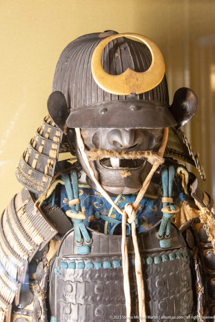 Close view of a samurai helmet, face guard, blue lacing, shoulder armor, and patterned textile.