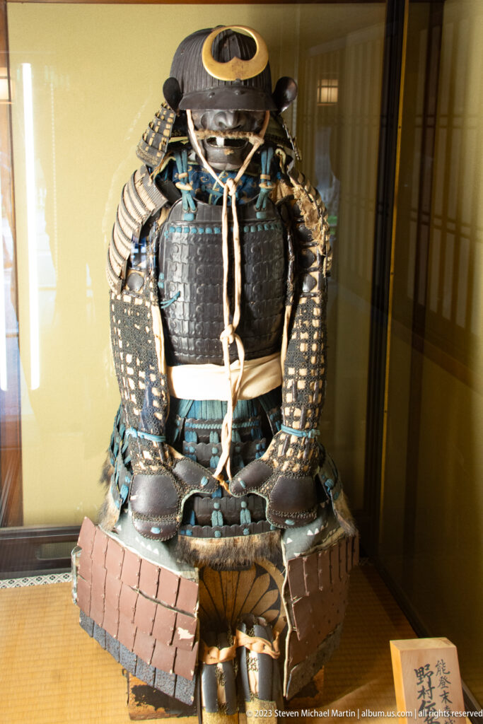 Front view of seated-form samurai armor with helmet, dark cuirass, blue-green lacing, and broad skirt plates.