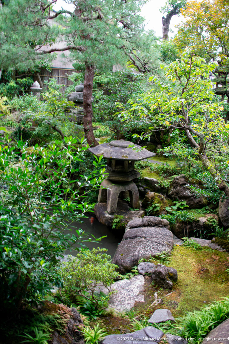 Mossy Japanese garden with stone lantern, stepping stones, shrubs, rocks, and layered green foliage.