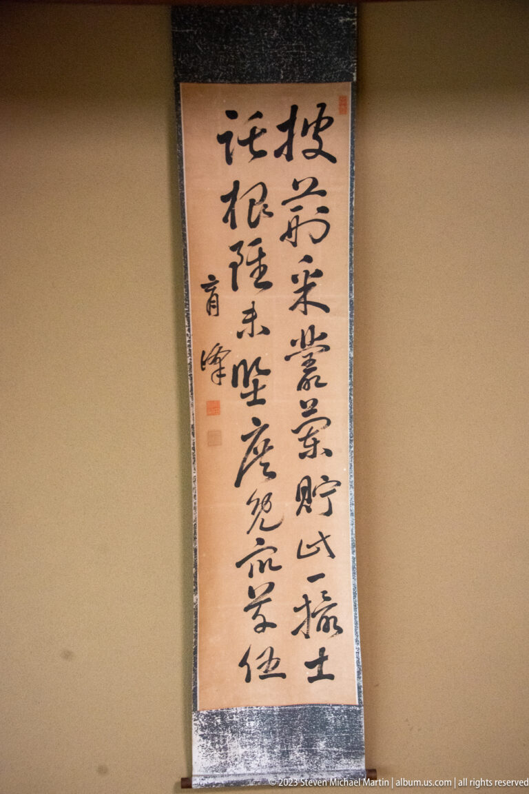 Vertical Japanese calligraphy scroll with bold black brushwork, textile mounting, and red seals on a plain wall.