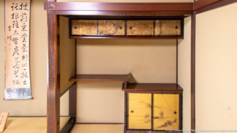 Tokonoma with calligraphy scroll, staggered shelves, gold-toned cabinet, timber framing, and tatami floor.