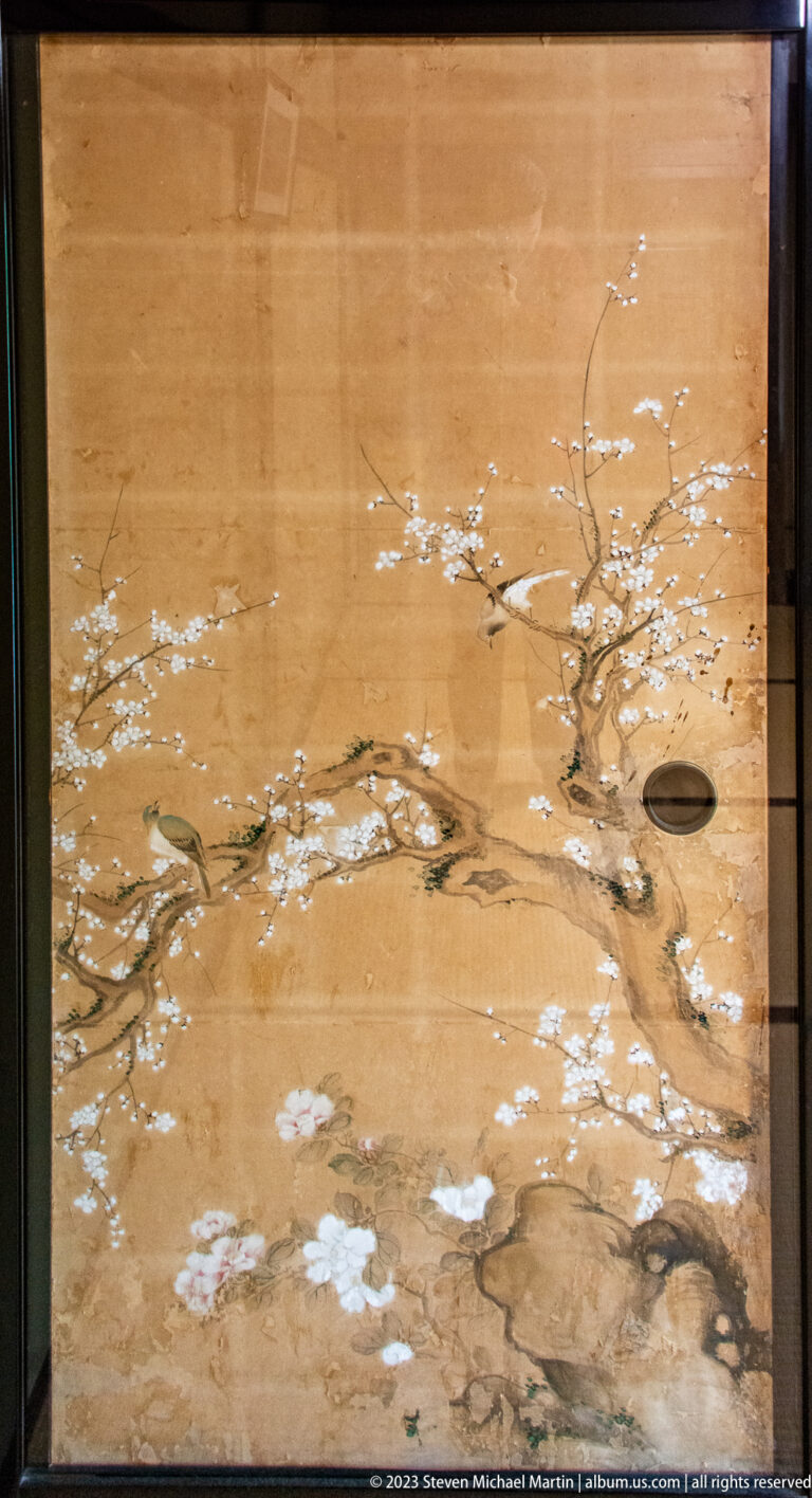 Gold-toned sliding-door panel painted with pale blossoms and branching twigs, with oval recessed pull at right.