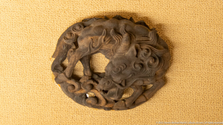 Round dark wooden carving with a lion-like head, curling foliage, and pierced openwork.