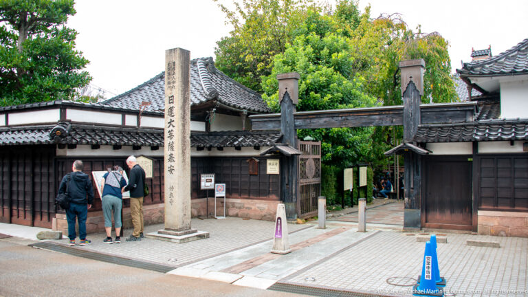 Wide view of Myōryū-ji’s street entrance with timber gates, white walls, tiled roofs, stone pillar, and visitors.