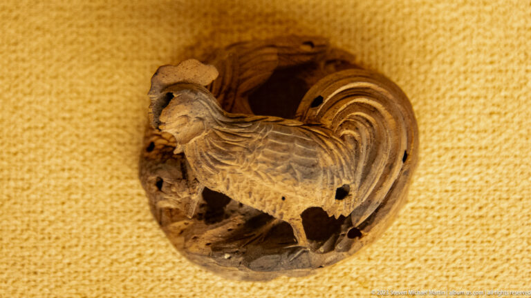 Round wooden openwork carving of a rooster with layered feathers, arched tail, comb, and beak.