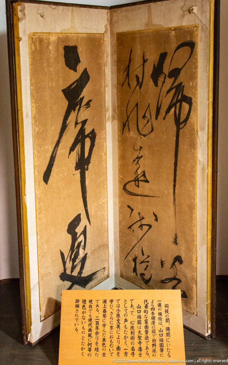 Gold-toned folding screen covered with large black calligraphy and finer vertical inscriptions, with label in front.