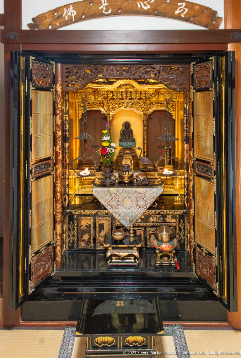 Open black lacquered Buddhist altar with gilded interior, seated Buddha, flowers, candles, and ritual vessels.