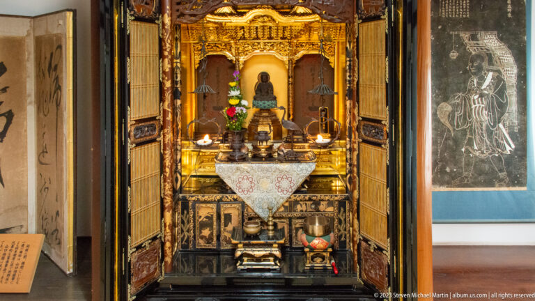 Full-height open butsudan with black doors, gilded shrine, seated Buddha, flowers, offerings, and lower compartments.