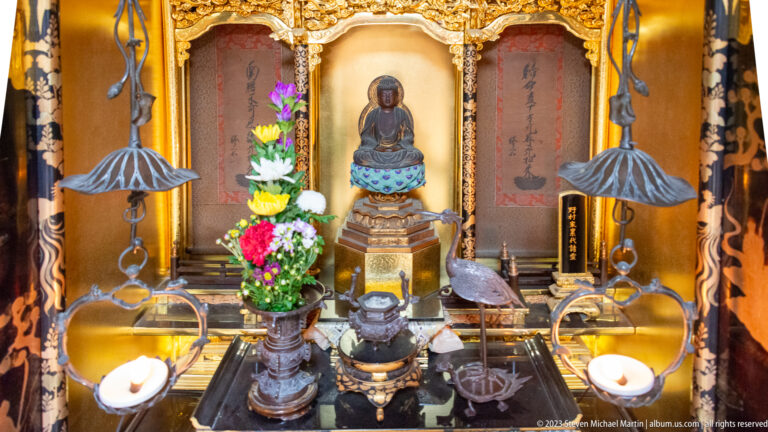 Close view inside a gilded Buddhist altar with seated Buddha, flowers, candles, incense burner, inscriptions, and hanging ornaments.