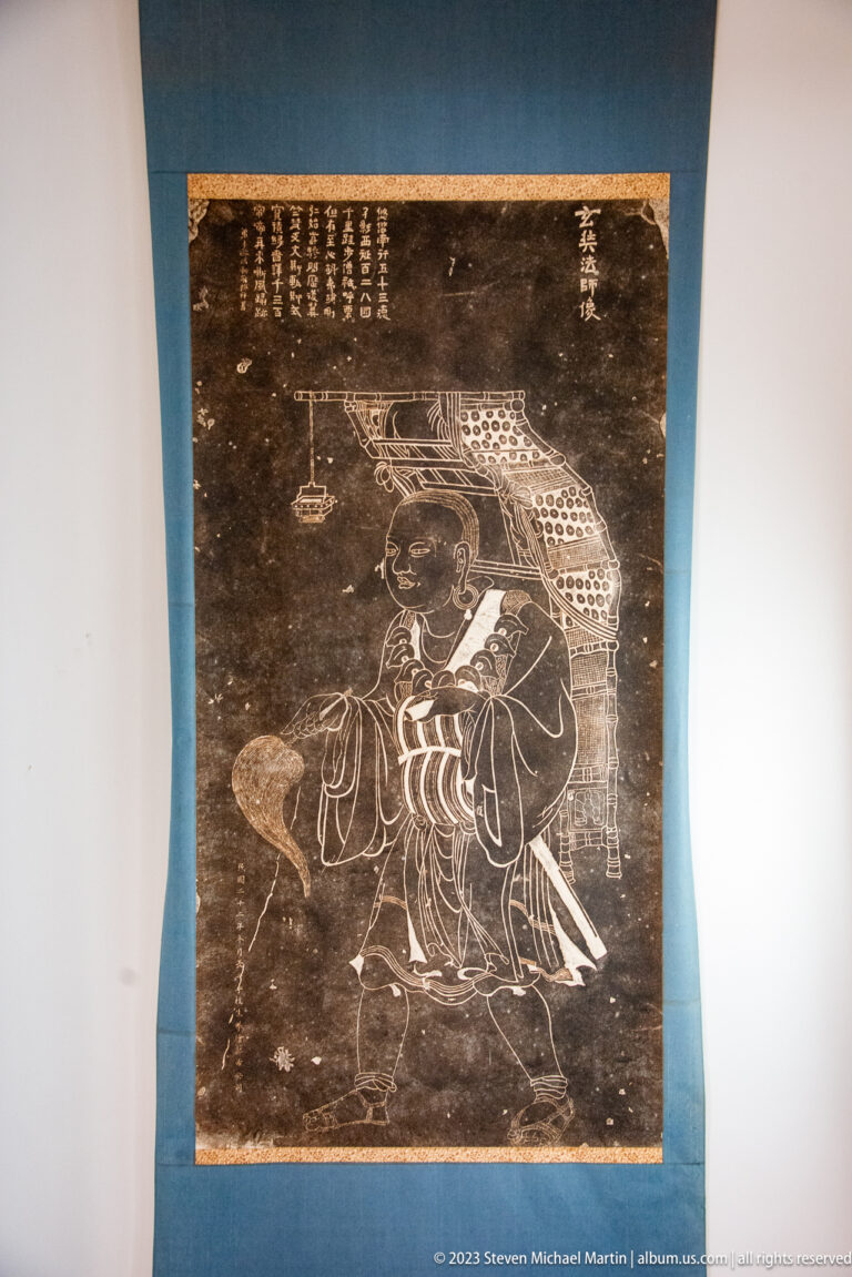 Dark monochrome hanging image with a robed figure, large spotted animal form above, and vertical inscriptions.