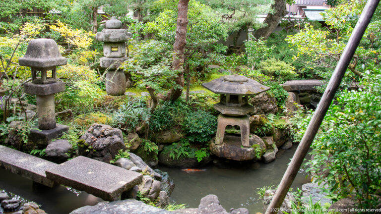 Garden pond with several stone lanterns, rocks, clipped shrubs, moss, and a narrow stone slab crossing the water.