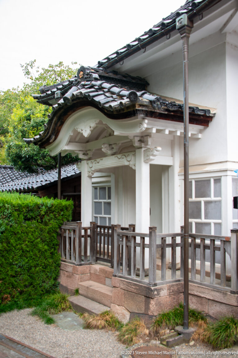 White-plastered temple building with curved tiled roof, wooden veranda, railing, and shrubs near Myōryū-ji.