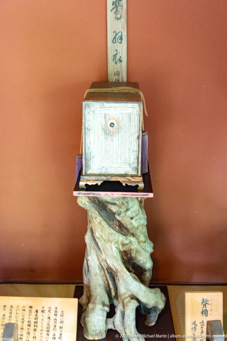 Small wooden chest on a natural-root pedestal with a hanging calligraphy scroll and exhibit labels.