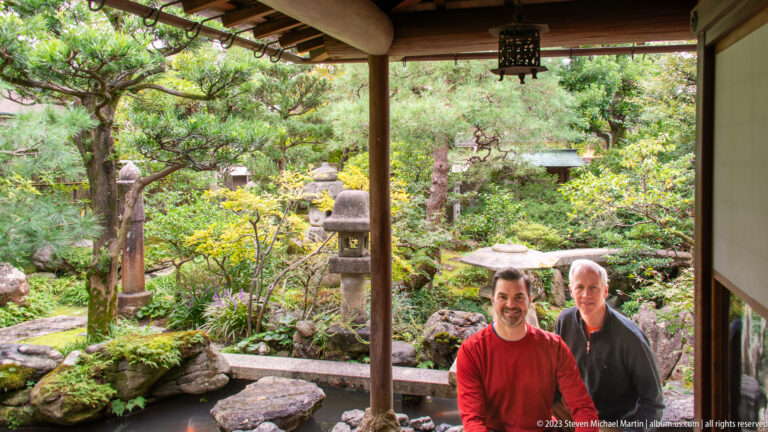 Two male visitors posing on a timber veranda with a pond, stone lanterns, and lush Japanese garden behind them.