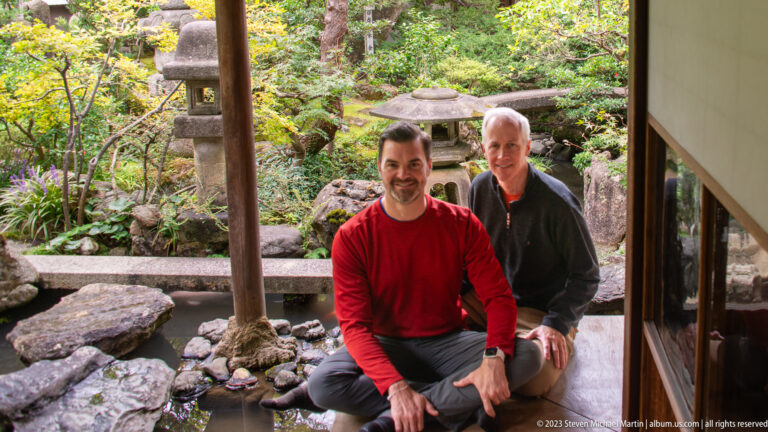Two male visitors seated on a wooden veranda beside a pond and stone-lantern garden.