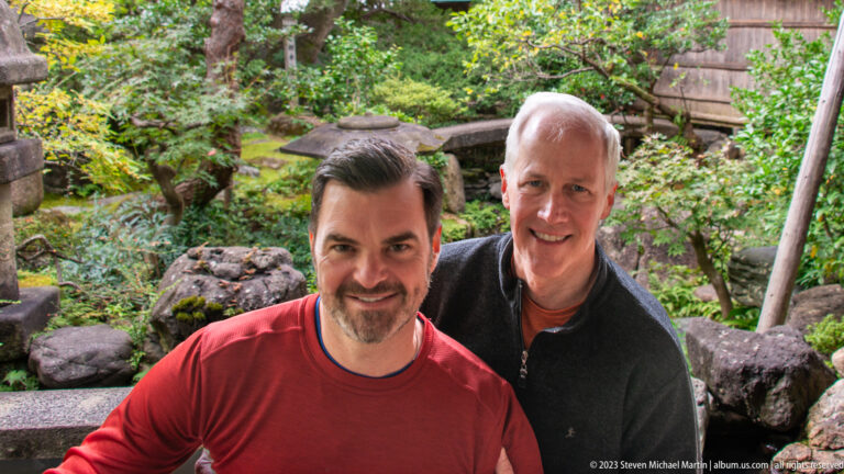Close portrait of two smiling male visitors with mossy rocks, shrubs, pond, and stone lantern behind them.