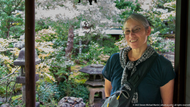 Female visitor standing beneath a timber veranda with mossy garden, rocks, and stone lantern behind her.