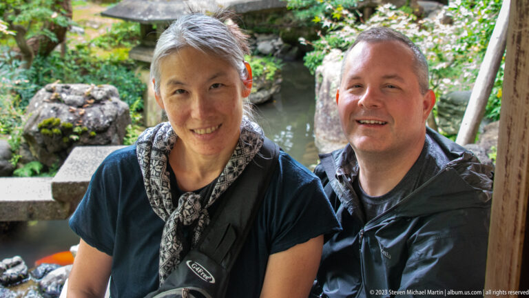 Two smiling visitors posing beside a garden pond with mossy rocks and stone lantern behind them.