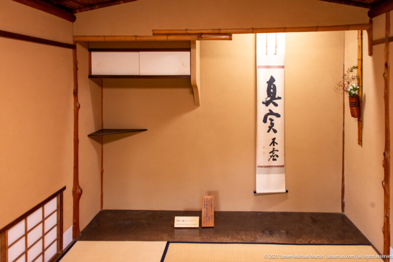 Compact tatami room with calligraphy scroll, built-in shelves, natural timber posts, and small floral accent.