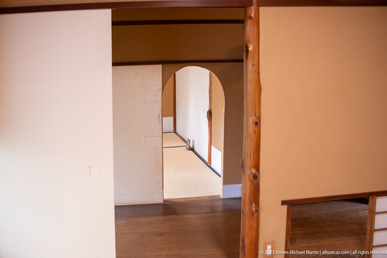 Rounded doorway in pale plaster wall connecting minimalist rooms with timber posts and polished wooden floors.