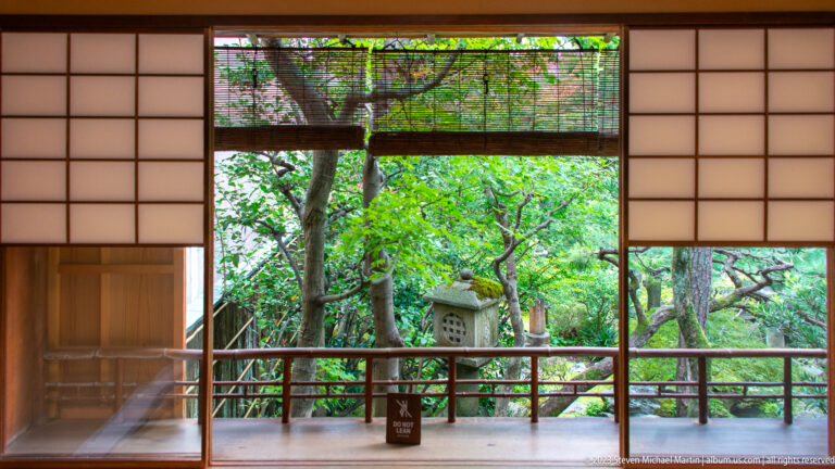 Garden-facing room with shoji panels, bamboo blind, veranda rail, tree, and stone lantern beyond.