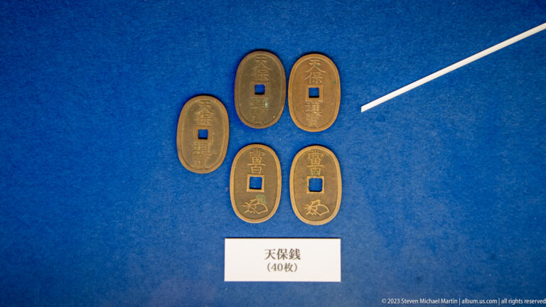 Five oval Tenpō coins with square central holes and raised inscriptions displayed on blue fabric.