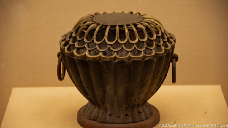 Dark ribbed armor-related object with looped lacing, domed top, and metal side rings on a display stand.