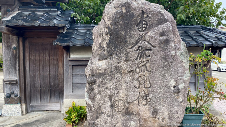 Vertically inscribed stone monument standing before a weathered wooden gate and tiled wall.