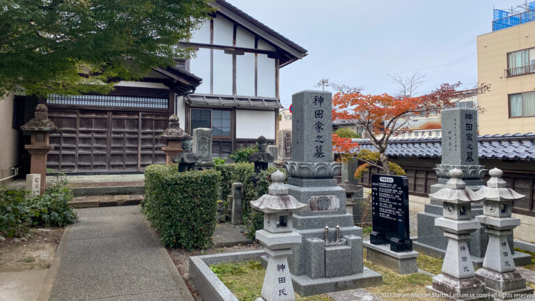 Temple courtyard with inscribed stone memorials, lanterns, grave markers, shrubs, and traditional buildings.